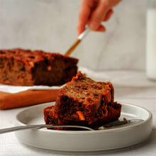 Two slices of dark banana bread rest on a white plate, with a hand and the loaf behind.