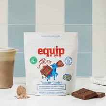 A white Equip Kids milk chocolate protein powder pouch stands beside a scoop of powder, chocolate squares, and a filled cup.