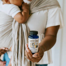 Blue supplement bottle labeled "Liquid Gold" held in hand by person wearing white shirt holding infant.