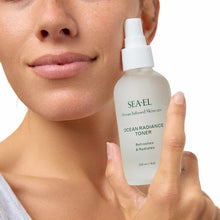 A woman holds a frosted bottle of Sea-El Ocean Radiance Toner with a white spray cap beside her cheek.