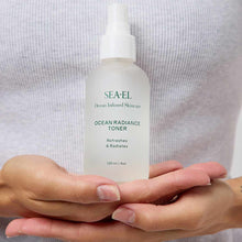 Two open hands hold a frosted glass spray bottle of Sea-El Ocean Radiance Toner against a gray ribbed top.