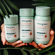 Three pale green bottles of True Grace Omega-3 Fish Oil supplement held in hands against green foliage background.