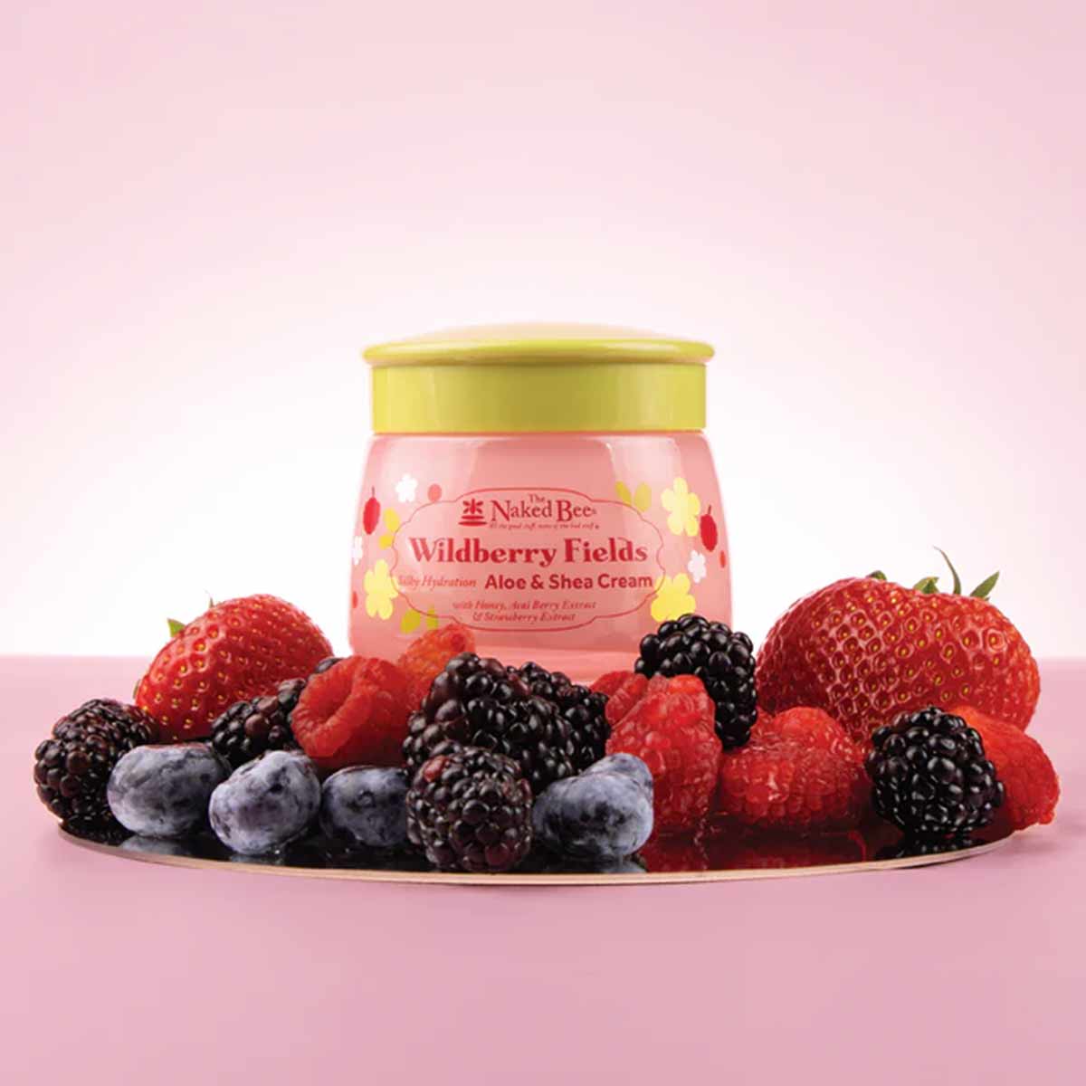Pink jar with yellow lid labeled "Wildberry Fields Aloe & Shea Cream" surrounded by strawberries, raspberries, blackberries, 