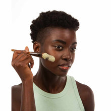 Woman applying tan clay with a wooden brush to her cheek.