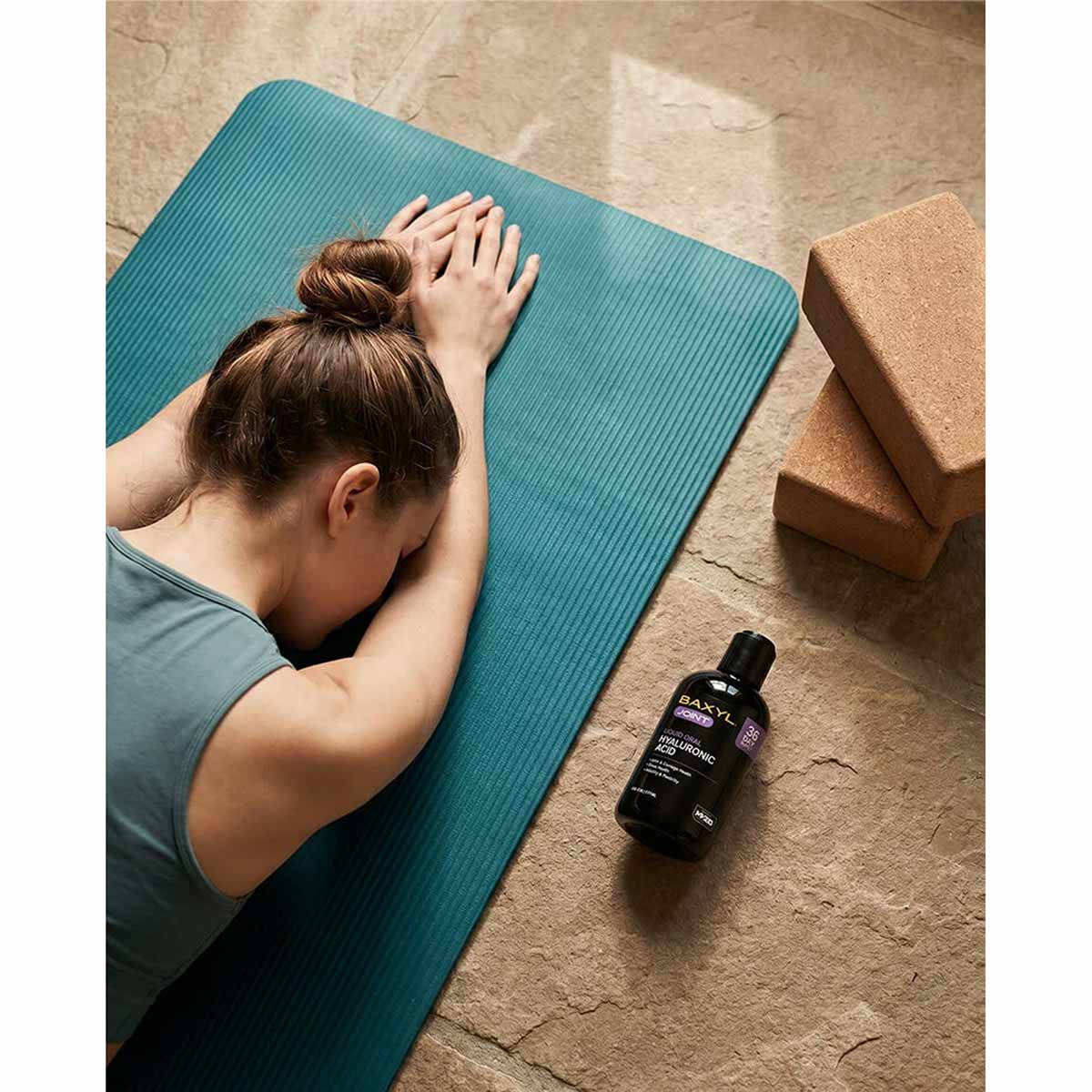 Black bottle of Baxyl Healthy Joints supplement on concrete floor next to blue yoga mat with person.