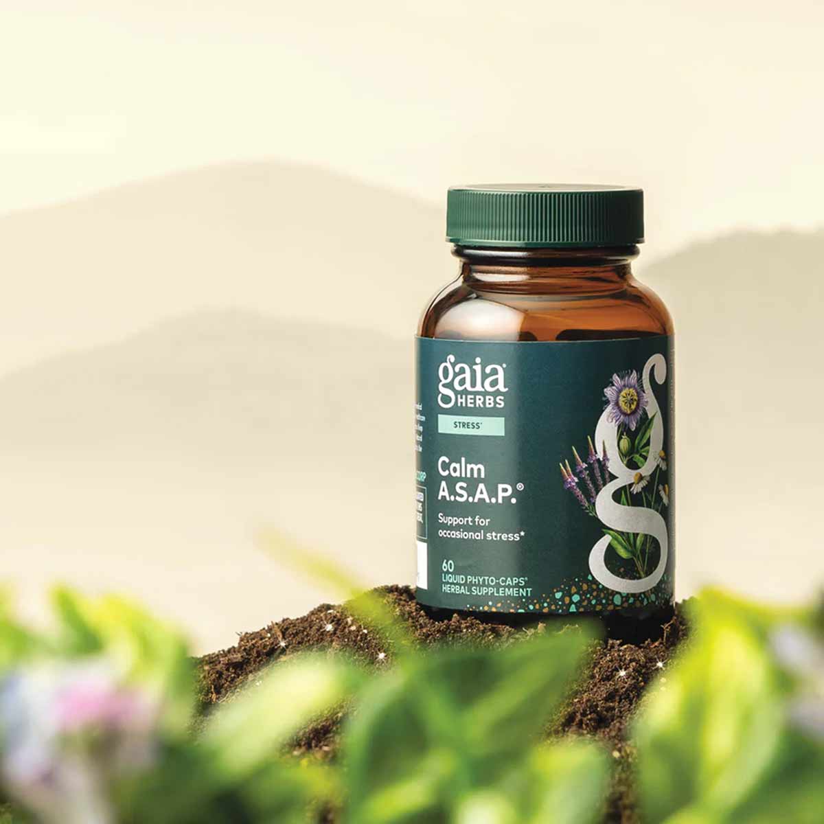 Gaia Herbs Calm A.S.A.P. amber glass bottle with green cap displayed on soil with blurred flowers and foliage background.
