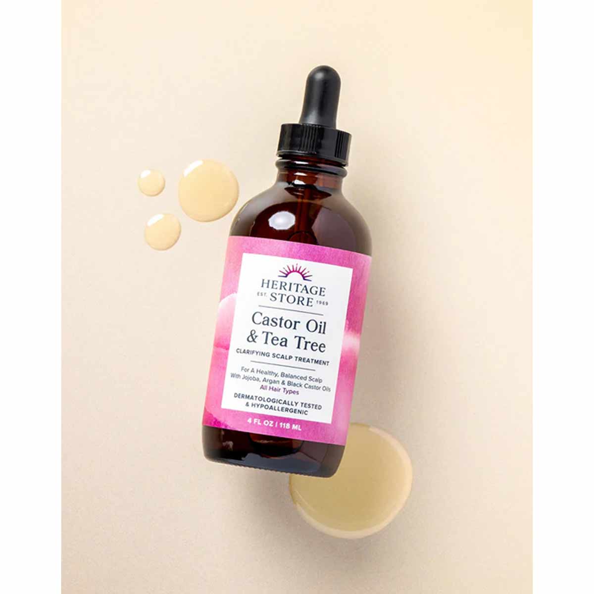 Dark amber glass bottle with black dropper top and pink label for Heritage Store Castor Oil & Tea Tree, surrounded by oil dro