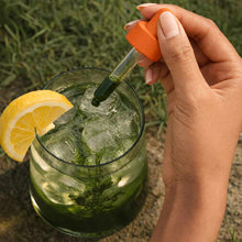 Hand holding dropper with orange cap dispensing liquid into glass of water with lemon slice and ice.