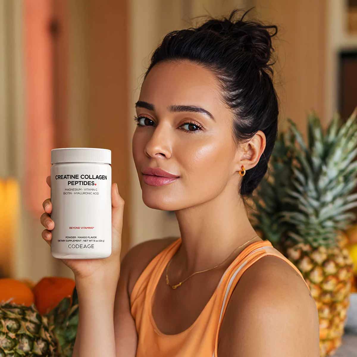 White cylindrical supplement container labeled "Creatine Collagen Peptides" held by woman in orange tank top.