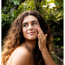 Person with long curly brown hair applying serum to cheek against leafy green background.