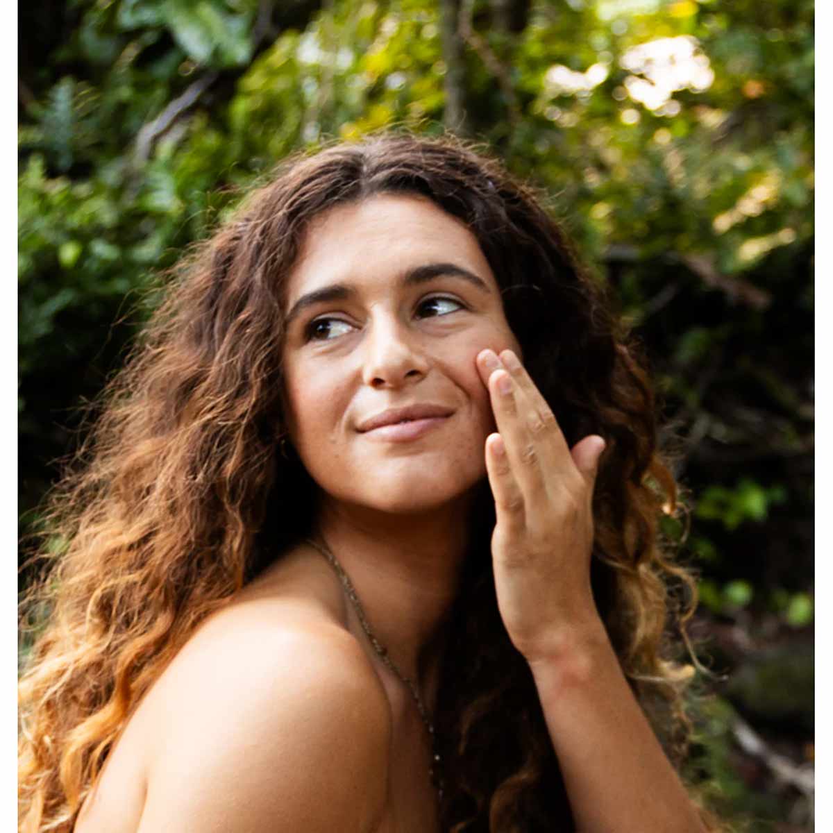 Person with long curly brown hair applying serum to cheek against leafy green background.