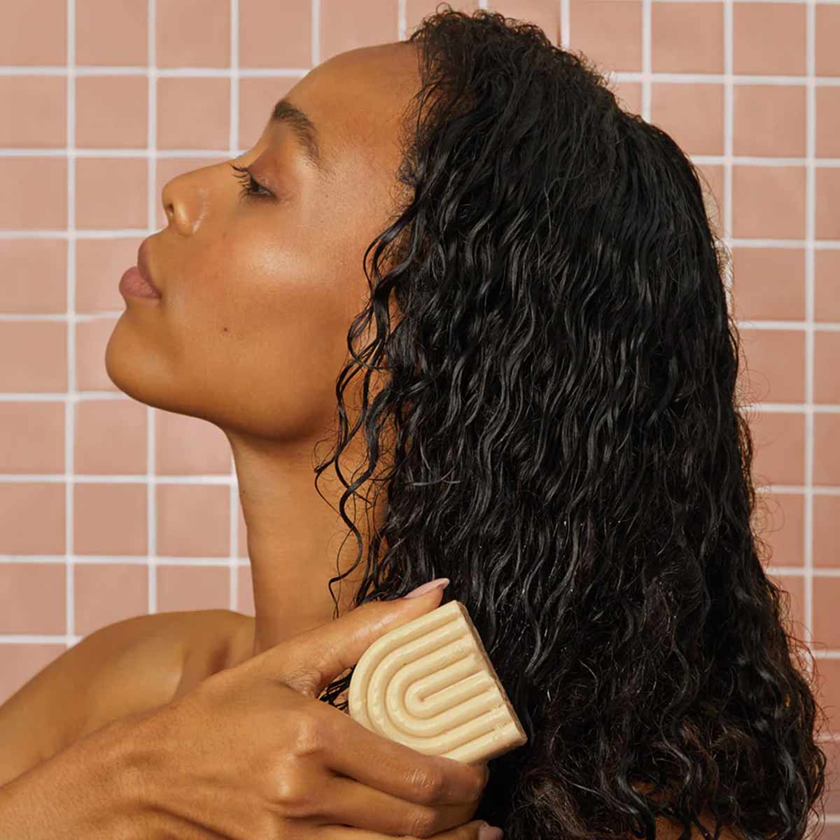 Person holding tan-colored solid conditioner bar against wet, dark curly hair in shower setting.