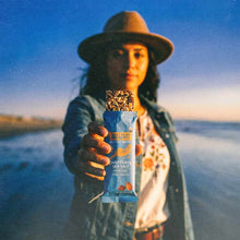 Chocolate sea salt energy bar in blue wrapper held by person wearing hat against water landscape background.