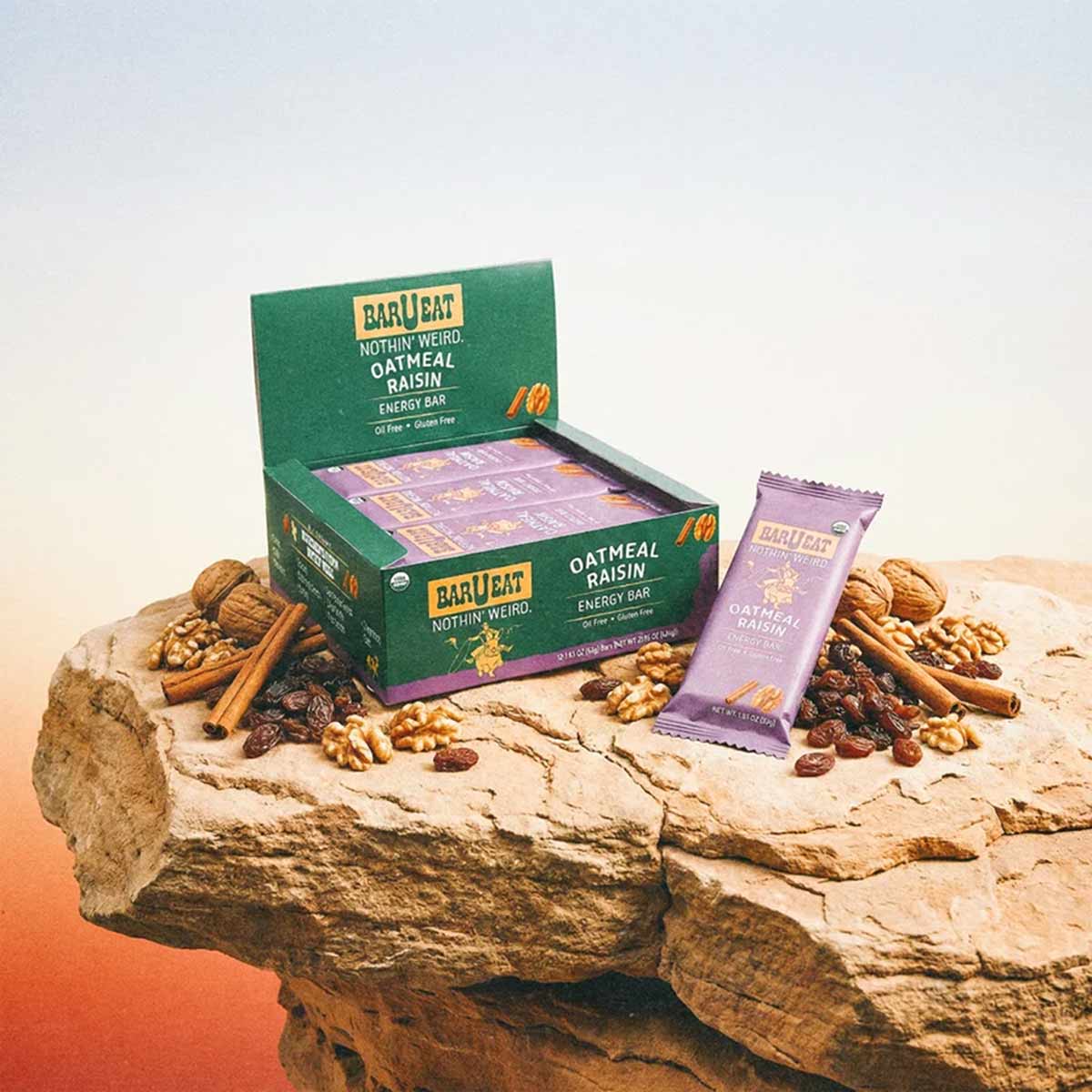 BAR U EAT oatmeal raisin energy bar box with individual wrapper and scattered walnuts and raisins on sand-colored rock.
