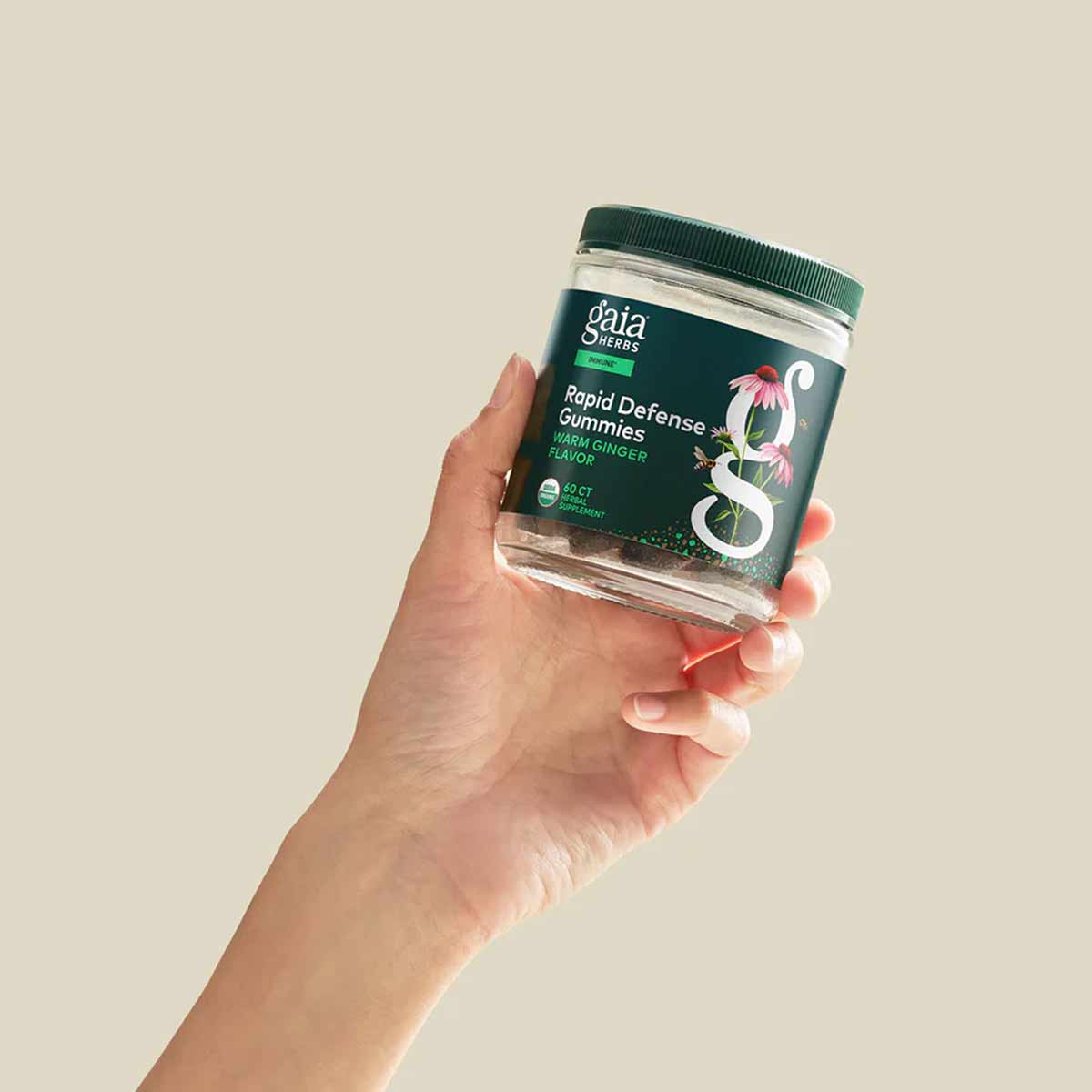 Hand holding a clear jar of Gaia Herbs Rapid Defense Gummies with dark green label and ginger flavor description.