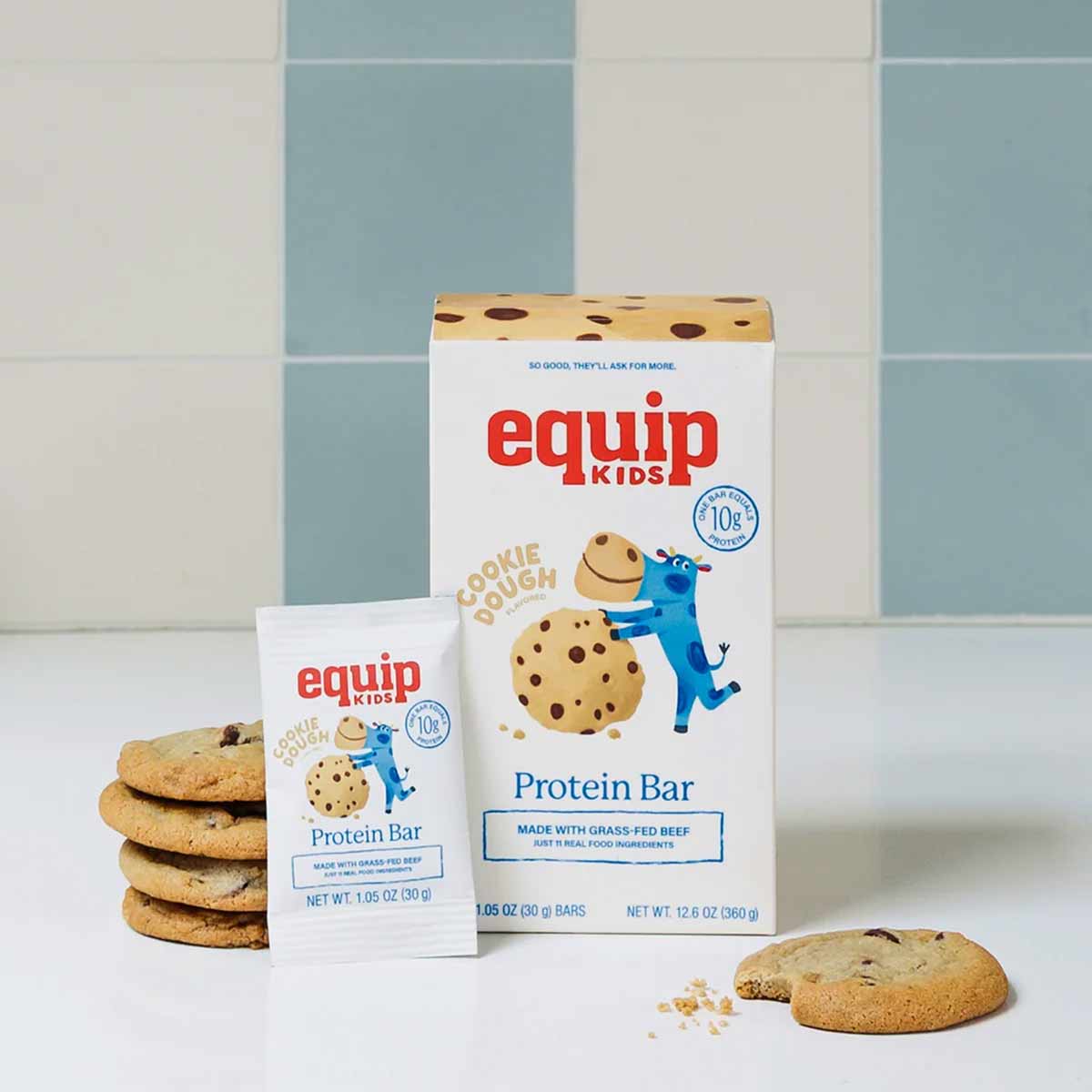A box and single wrapper of Equip Kids cookie dough protein bars sit beside stacked chocolate chip cookies against blue and white tiles.