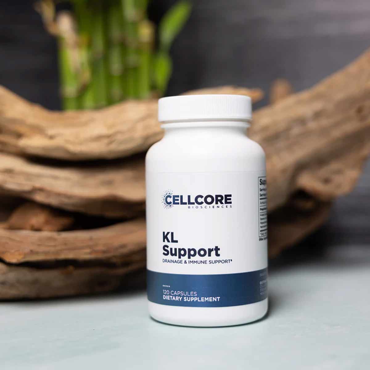 White bottle of CellCore KL Support dietary supplement capsules with blue label, displayed with driftwood and green plant.
