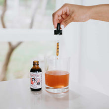 Hand holding dropper dispensing amber liquid from Lactivist bottle into clear glass of liquid.