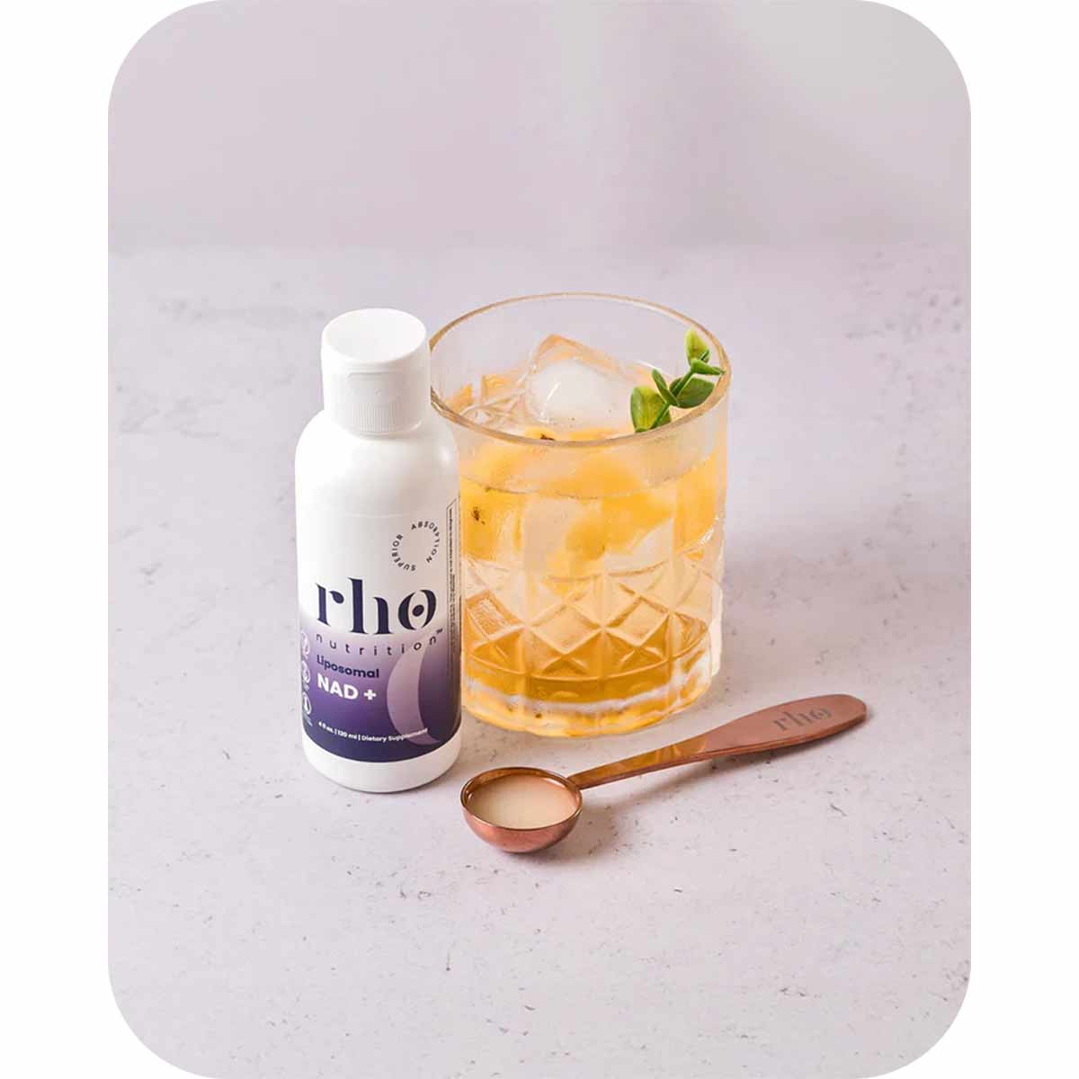 Rho Nutrition Liposomal NAD+ bottle next to a glass of amber liquid with ice and mint, with a measuring spoon.