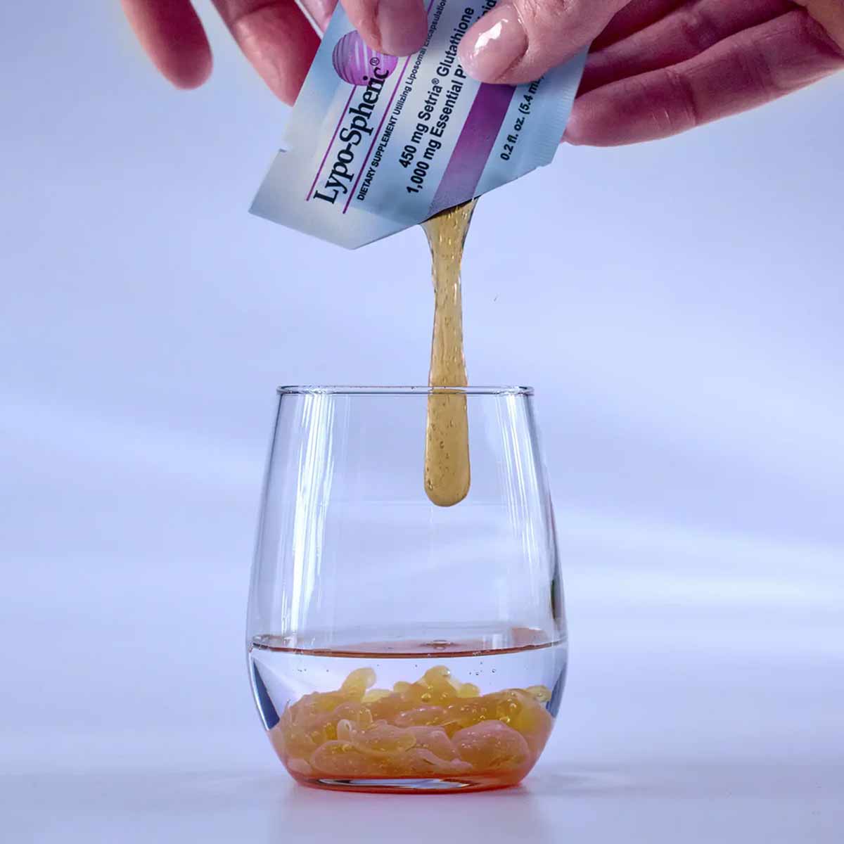 Hand squeezing Lypo-Spheric Glutathione sachet, dispensing golden liquid into glass.