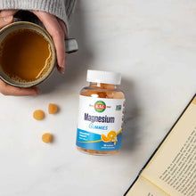 Bottle of Kal Magnesium Gummies with orange gummies displayed nearby, next to a mug of tea and open book.