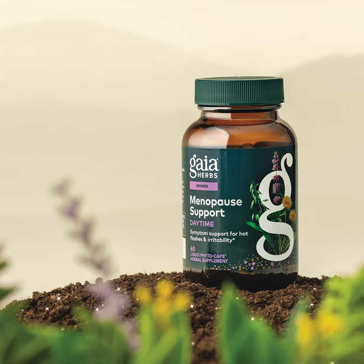 Gaia Herbs Menopause Support Daytime bottle with green cap on soil with plants and flowers.