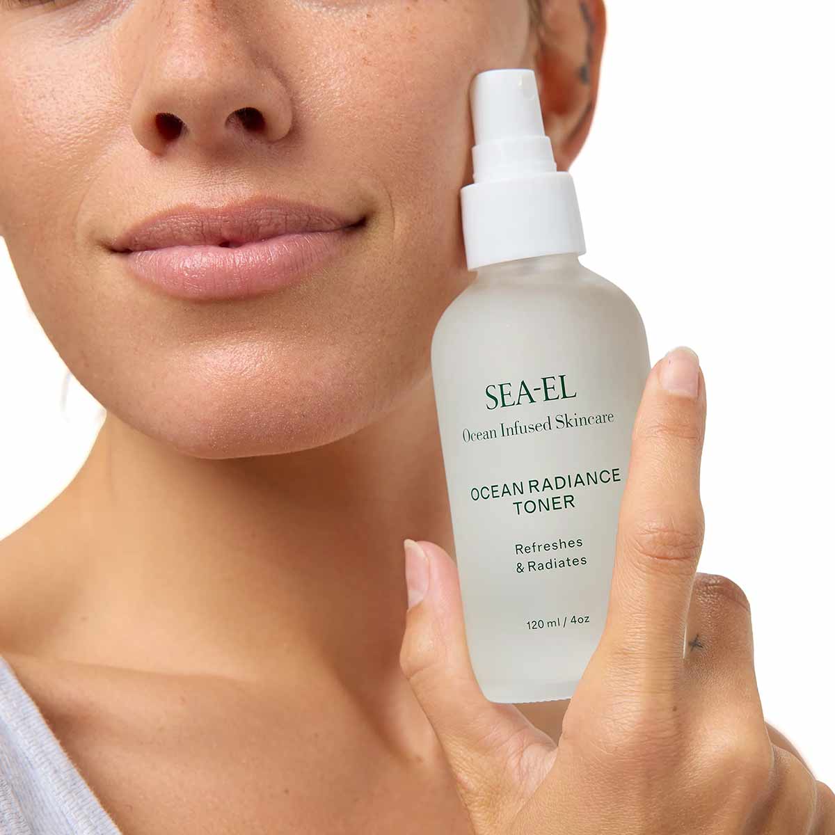A woman holds a frosted bottle of Sea-El Ocean Radiance Toner with a white spray cap beside her cheek.