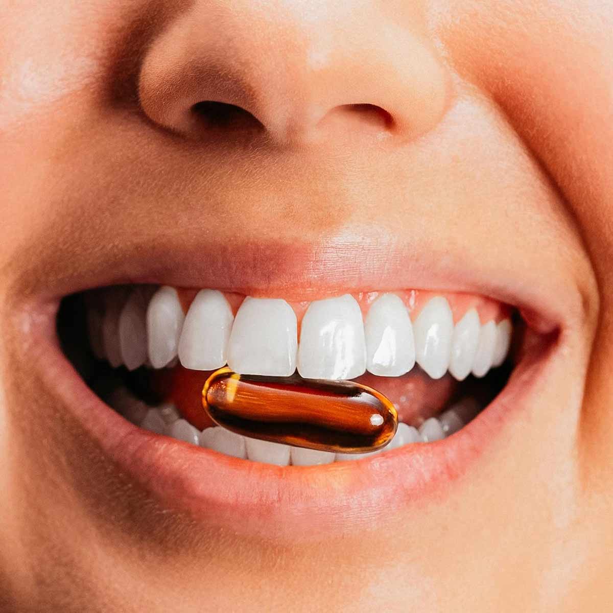 Person's mouth with white teeth holding an amber-colored fish oil capsule between the lips.