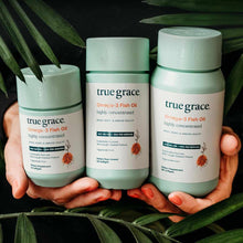 Three True Grace Omega-3 Fish Oil supplement bottles in pale green containers held in hands against a dark background with gr