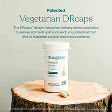 True Grace One Daily probiotic bottle with white cap and two vegetarian capsules on a wood surface.