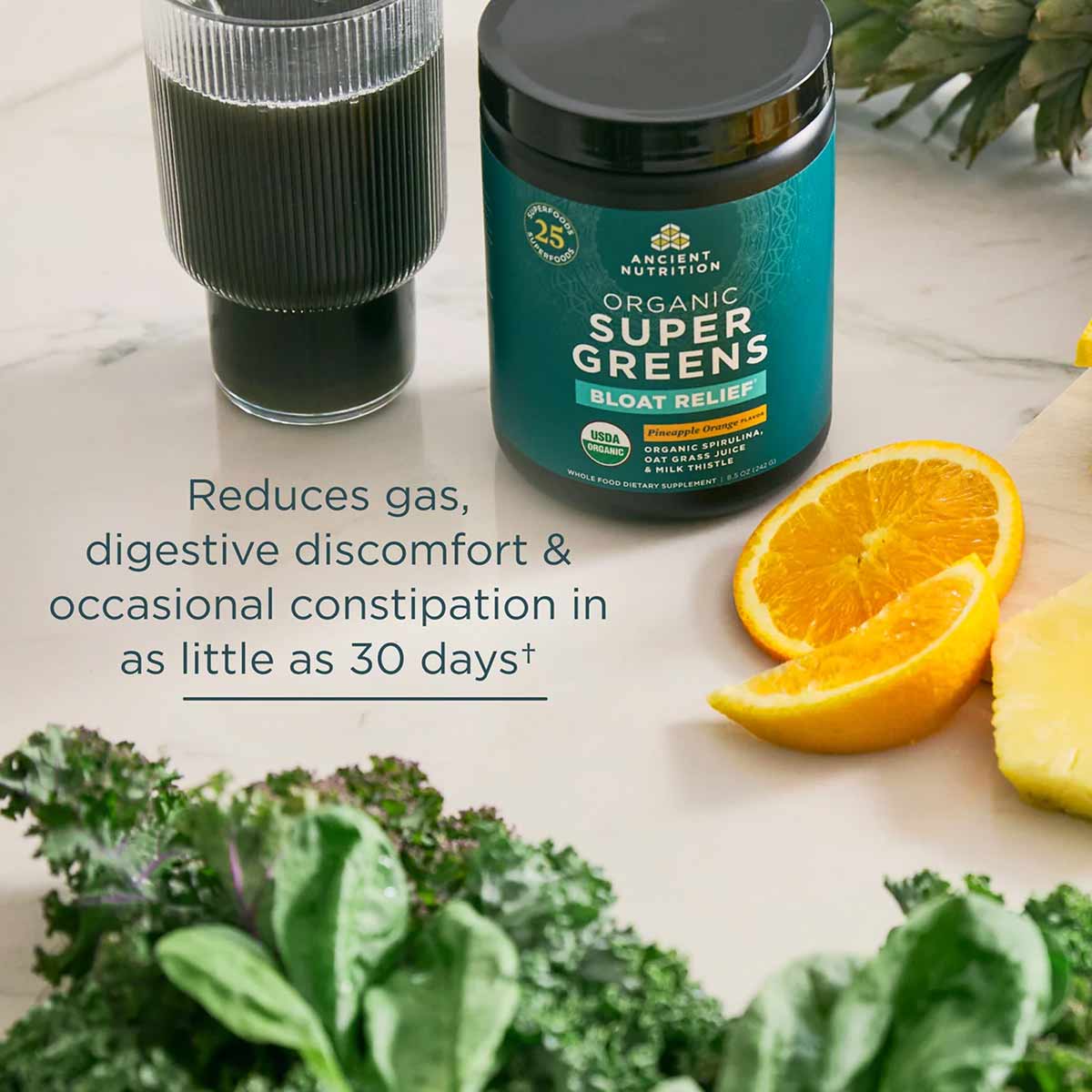 Teal container of Ancient Nutrition Organic SuperGreens + Bloat Relief powder with glass of green beverage, orange slices, an