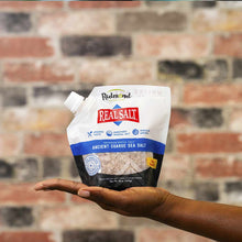White pouch of Real Salt coarse sea salt held in an open hand against a brick wall background.