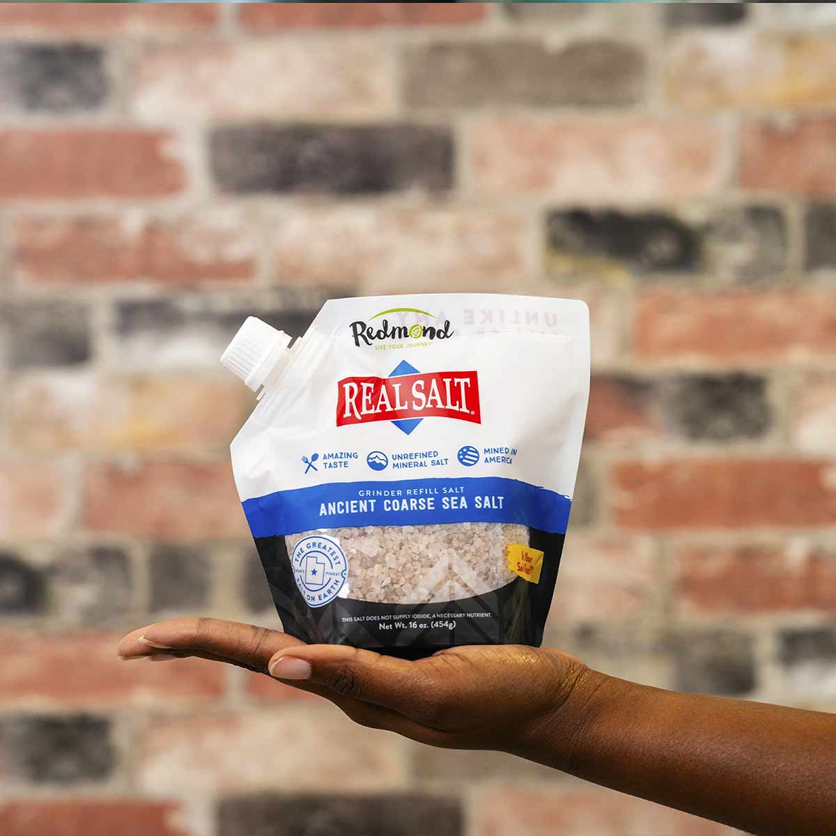White pouch of Real Salt coarse sea salt held in an open hand against a brick wall background.