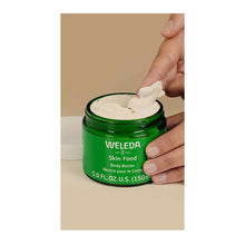 Hand scooping cream from open green jar labeled Weleda Skin Food Body Butter, 5.0 FL OZ.