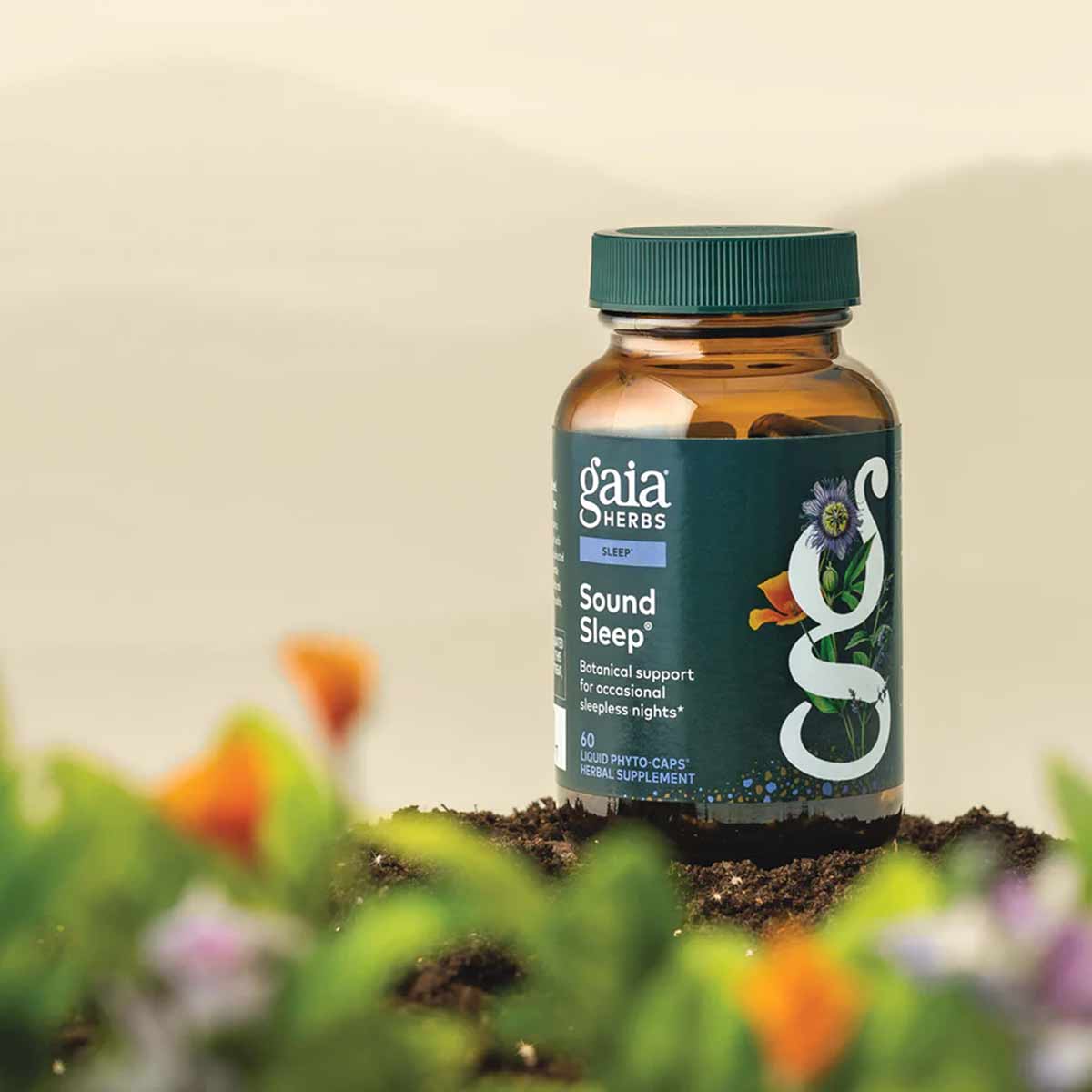Gaia Herbs Sound Sleep bottle with teal cap sitting on soil surrounded by blurred flowers and foliage.