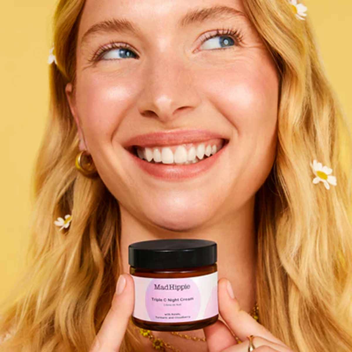 Woman with reddish-blonde hair holding a pink-labeled Mad Hippie Triple C Night Cream jar with black lid against yellow backg