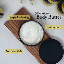Open jar of white body butter on wooden board with black jar lid nearby.